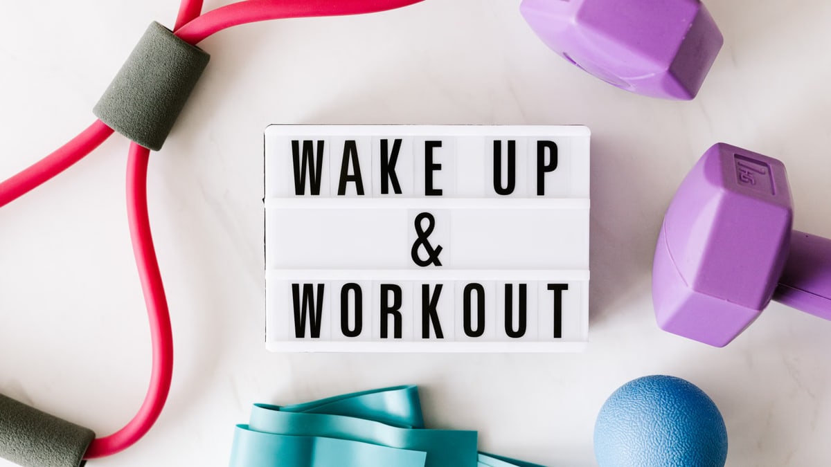Top view closeup composition of wake up and workout inscription on light box surface among expanders and dumbbells and massage ball on white background